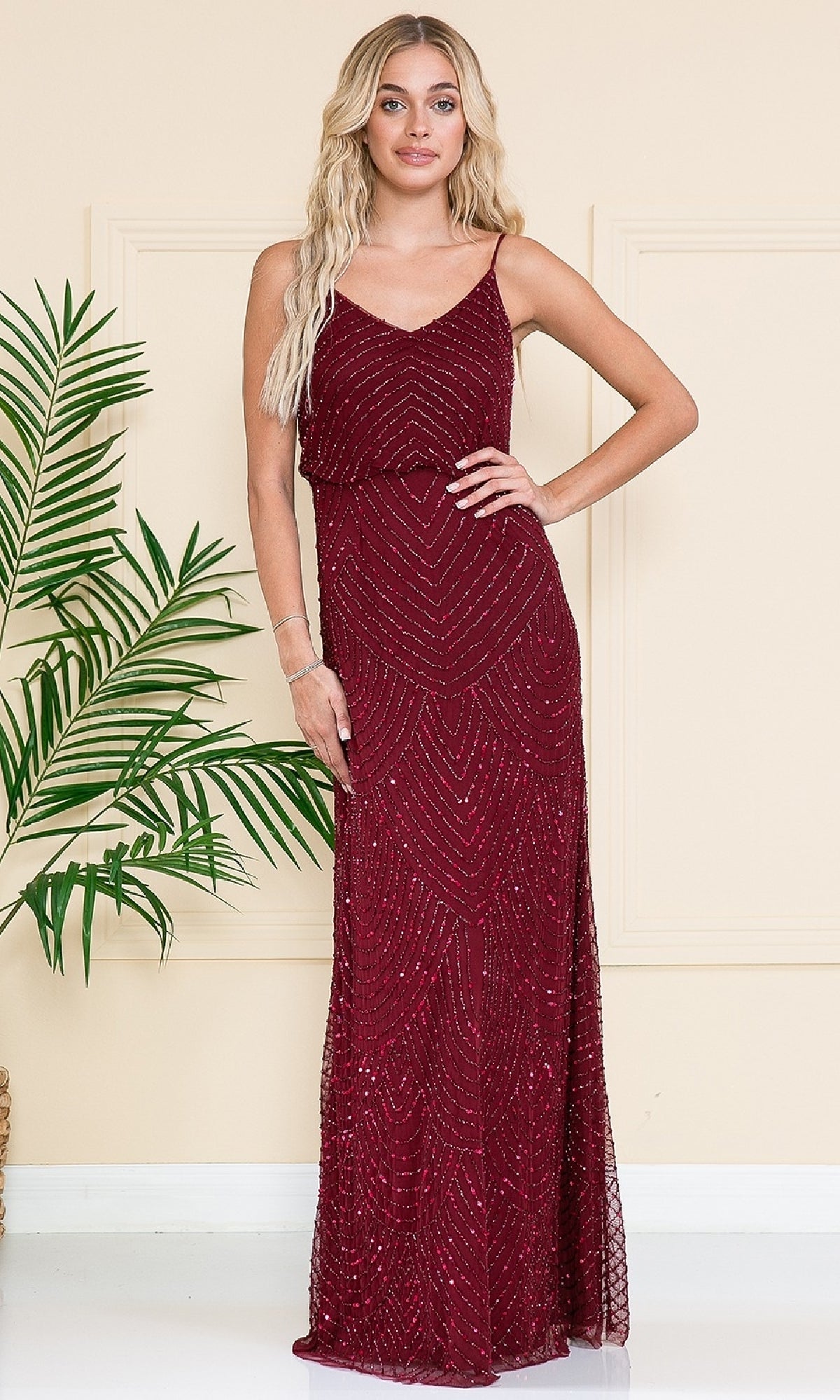 A woman in the Amelia Couture IN001 Sequin Blouson Evening Gown stands indoors beside a green potted plant against a beige wall.